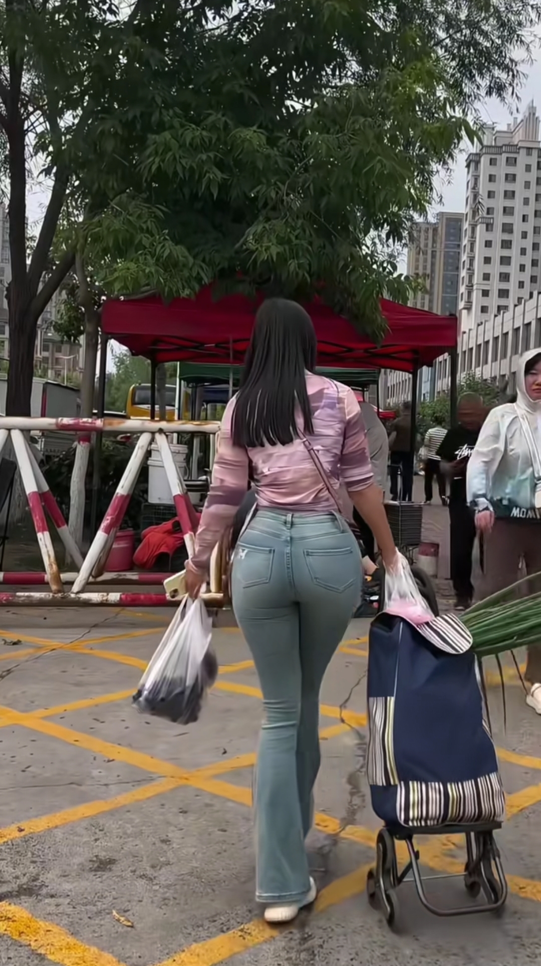 街头偶遇，她穿着时髦，粉紫上衣配喇叭牛仔裤，拖着小推车，拎着购物袋，背影都藏着对生活的热爱，平凡日常也能这么有范儿，咱普通人的鲜活劲儿，就藏在这烟火气里啦～